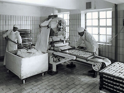 Schwarz-weiß Foto bei dem 2 Männer an Maschinen stehen und Käse zubereiten.
