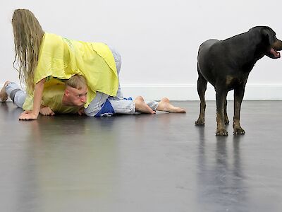 Foto: Performances for Pets, Performance for Orpheus, 2020 -  die Künstler Krõõt Juurak und Alex Bailey auf dem Boden liegend, davor steht ein großer schwarzen Hund