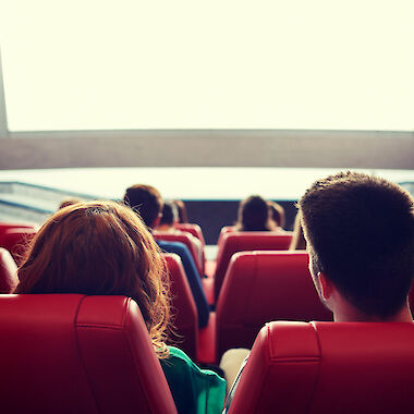 Kinobesucherinnen und Besucher von hinten auf roten Kinostühlen vor einer Leinwand.