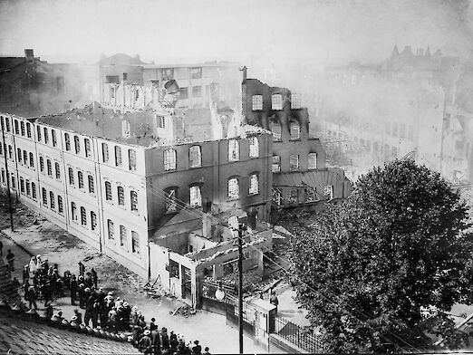 Der große Brand im Opelwerk von 1911. Eine Menschenmenge versammelt sich vor den ausgebrannten Fabrikhallen.