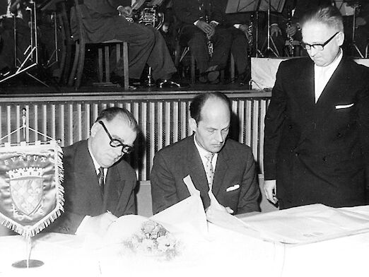 3 Männer, 2 sitzend, einer stehend, beim Unterzeichnen der er Verschwisterungsurkunde zwischen Rüsselsheim und Evreux.