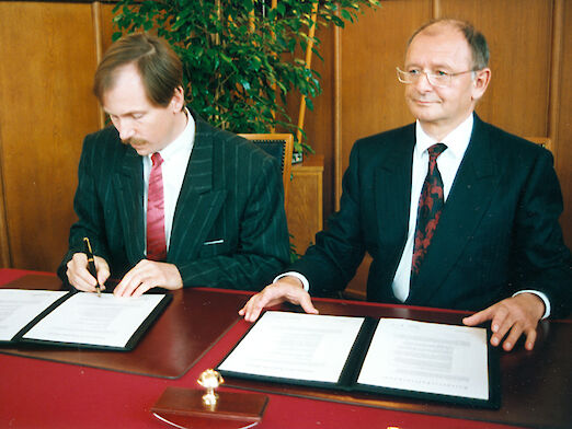 2 Männer, die an einem Tisch sitzen und den Partnerschaftsvertrag zwischen Rüsselsheim und Kecskemét unterzeichnen.