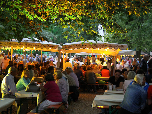 Gäste, die am Abend im Weindorf des Verna-Parks an Tischen und Bänken sitzen.