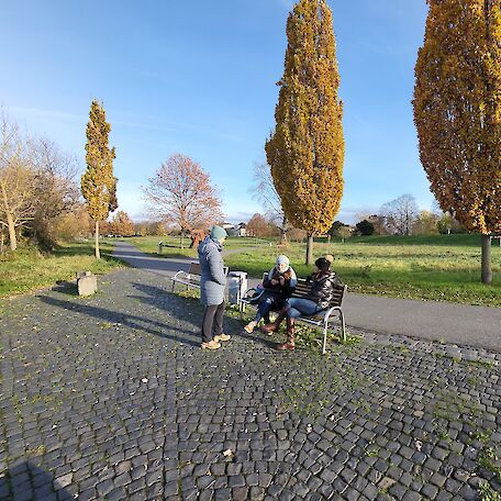 3 Frauen, eine steht und 2 sitzen auf einer Bank. Im Hintergrund Wiesen und Bäume im Herbst.