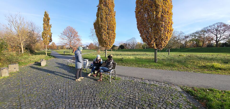 3 Frauen, eine steht und 2 sitzen auf einer Bank. Im Hintergrund Wiesen und Bäume im Herbst.
