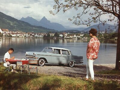 Ein Opel parkt an einem See, davor sind ein sitzender Mann und eine Frau zu sehen.